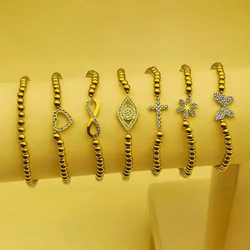 A collection of seven gold-beaded bracelets with various charm designs, including a heart, infinity symbol, evil eye, cross, flower, and butterfly, some adorned with clear crystals.